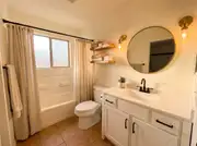 Refreshed small bathroom with a round brass mirror, black vanity pulls, warm sconces, wood shelves, and a linen curtain