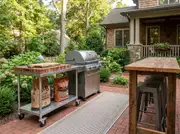 Rolling stainless prep cart holding bagged charcoal and wood pellets next to a gas grill on a brick patio