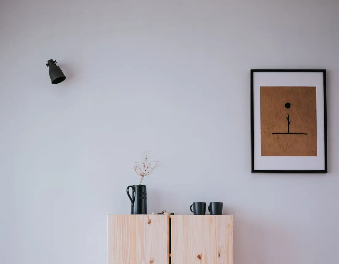 A ceramic pitcher and dried flower stem on a light wood cabinet beneath a framed line-art print
