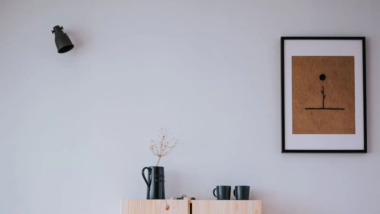 A ceramic pitcher and dried flower stem on a light wood cabinet beneath a framed line-art print