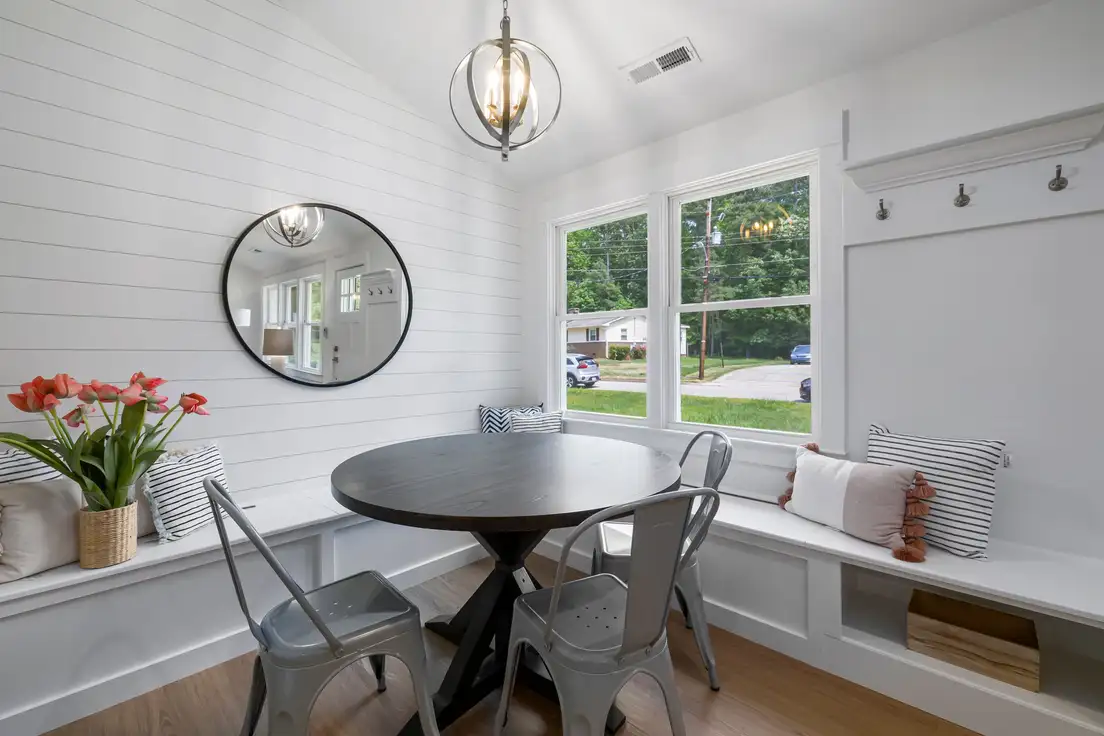 A round pedestal dining table in a built-in window banquette nook with cushions, metal chairs, and a round mirror on a white shiplap wall