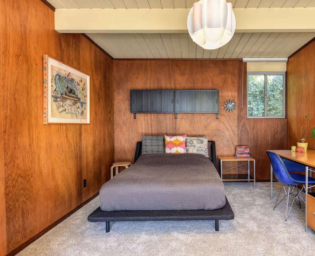 A paneled bedroom in a mid-century ranch with an exposed beam ceiling, a low platform bed, and a white pendant light