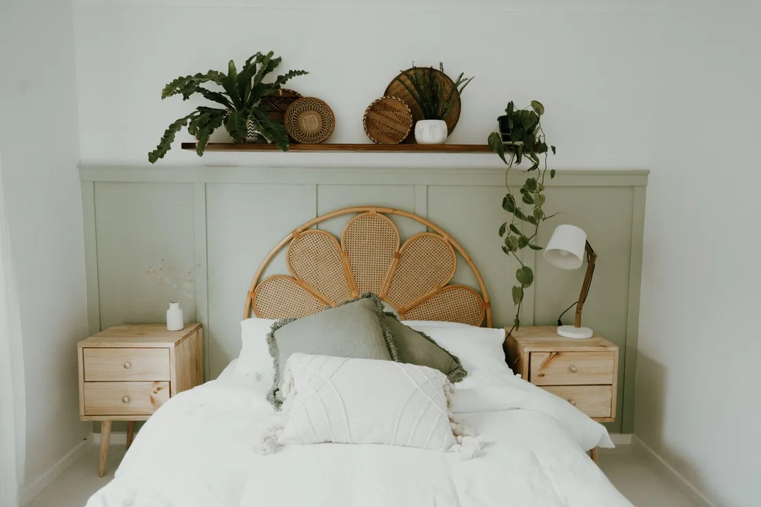 A rattan cane headboard with layered white and green pillows on a bed against a sage green wall