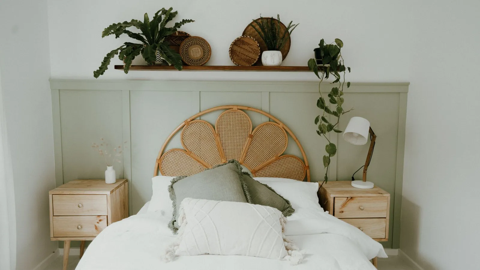 A rattan cane headboard with layered white and green pillows on a bed against a sage green wall