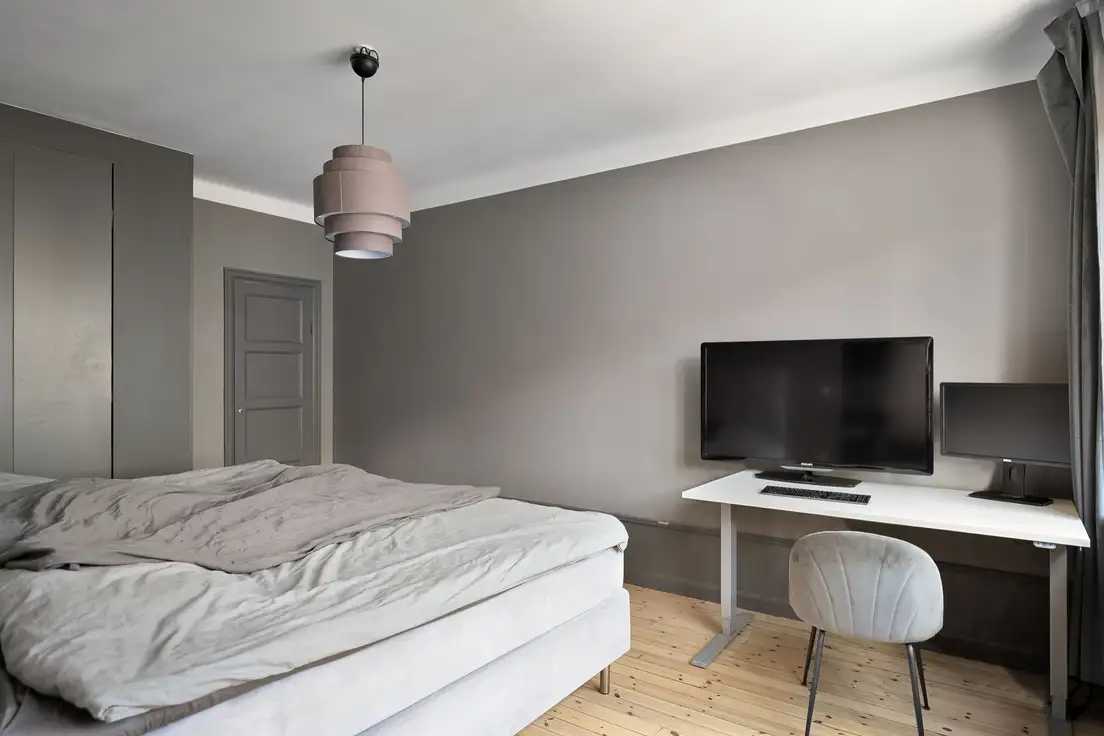 A gray bedroom with a white desk, a monitor, and an upholstered chair placed a few feet from the bed