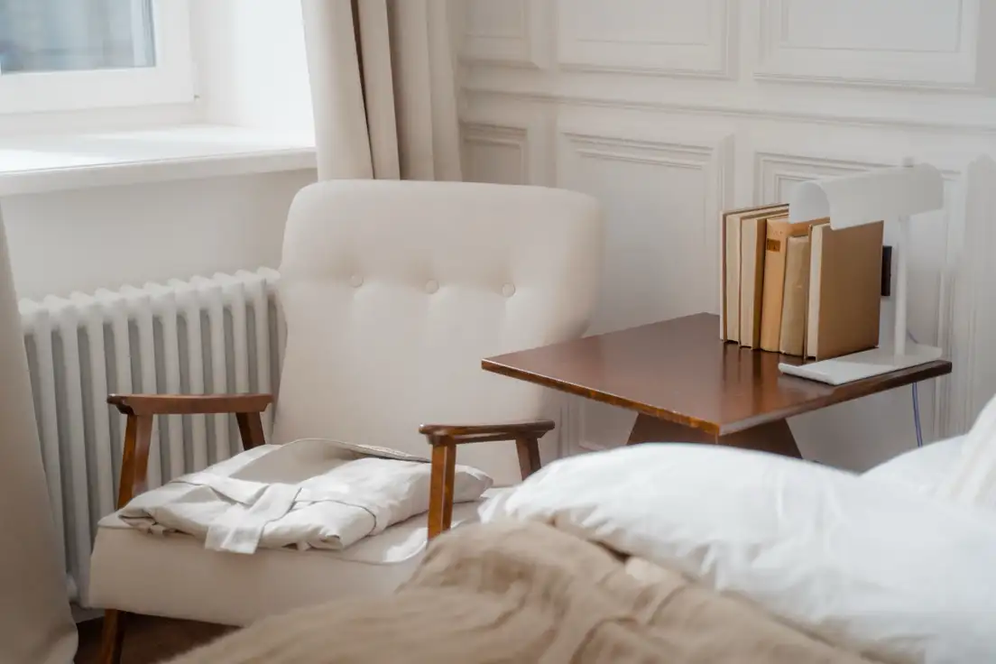 A cream accent chair with a robe draped over it beside a small wood table with books and a white lamp next to a bed