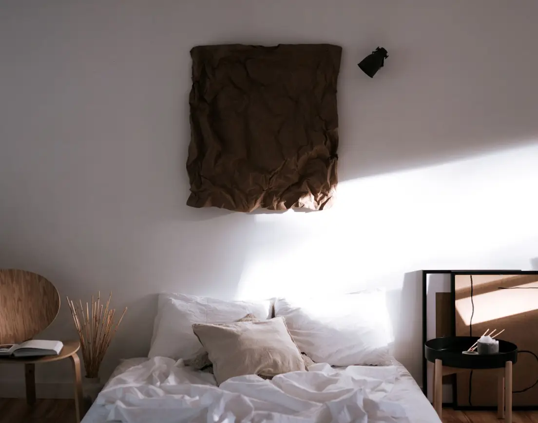 A warm minimalist bedroom with linen bedding, a textured brown wall hanging, and morning light