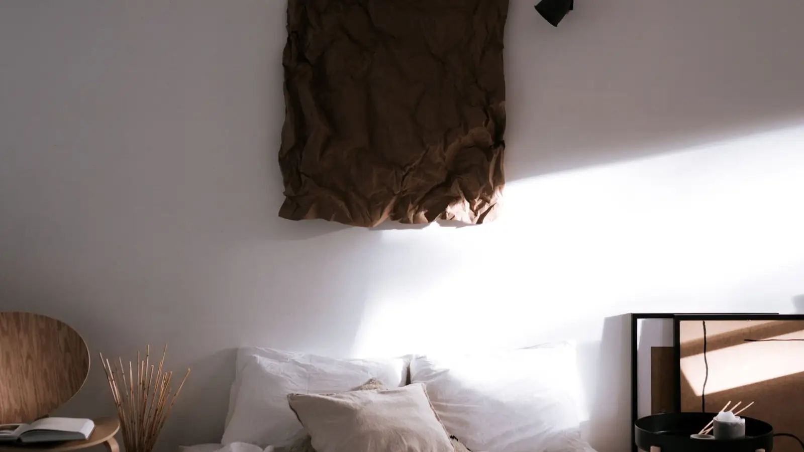 A warm minimalist bedroom with linen bedding, a textured brown wall hanging, and morning light