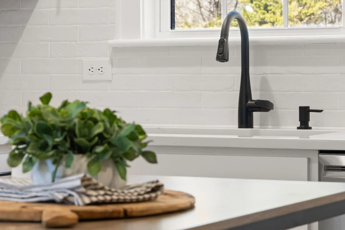 A black matte kitchen faucet against a white brick backsplash with a potted plant on the counter