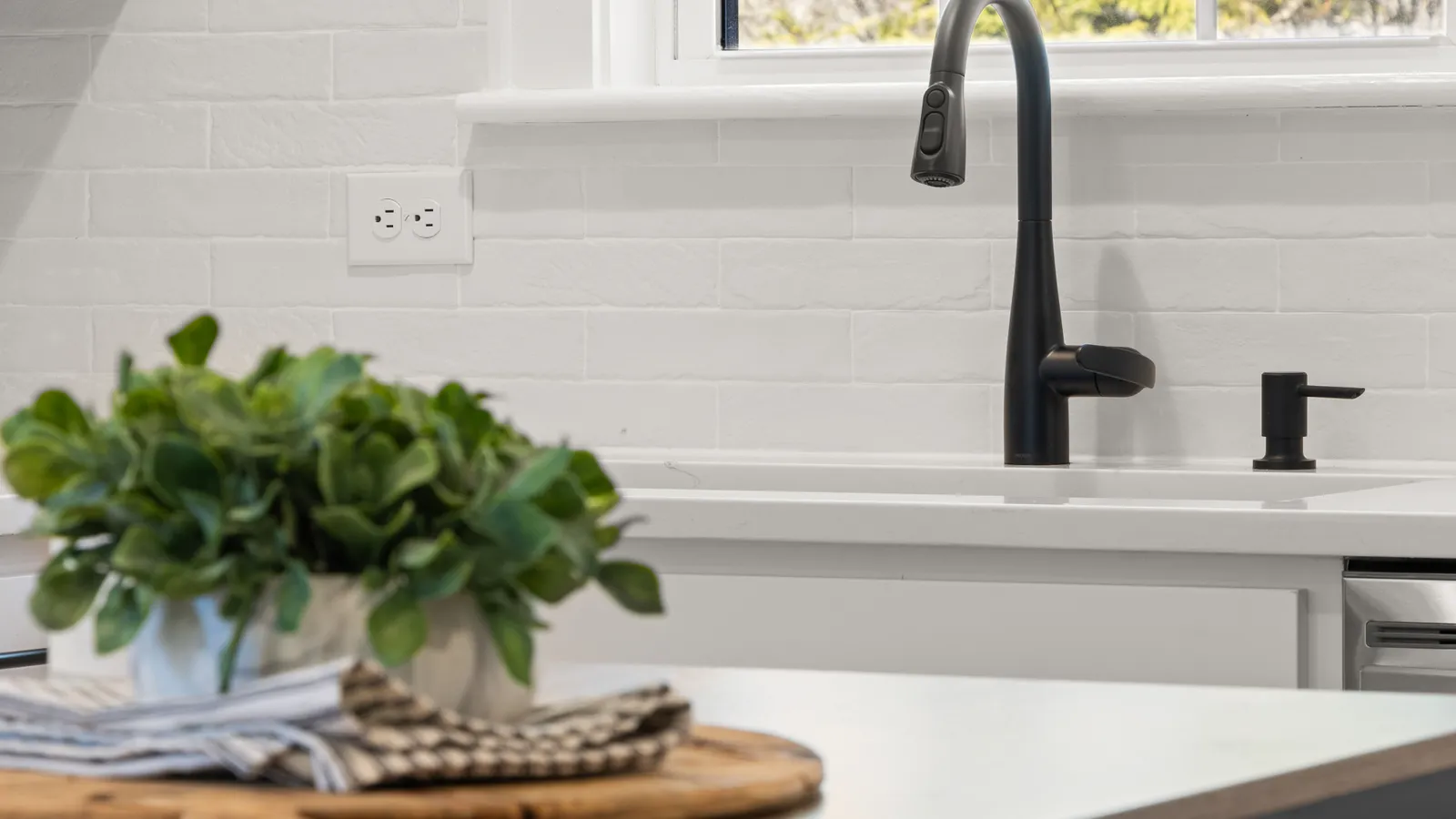 A black matte kitchen faucet against a white brick backsplash with a potted plant on the counter
