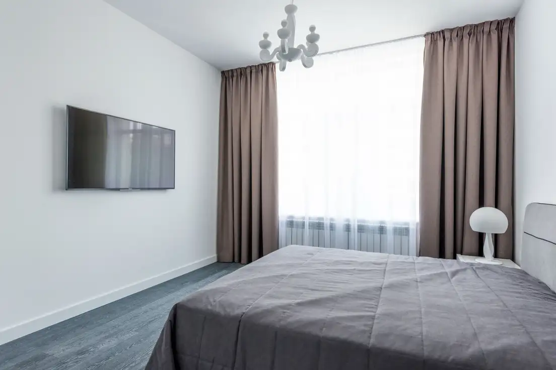 A bedroom with floor-length taupe blackout curtains over sheer panels, a gray bedspread, and a wall-mounted TV