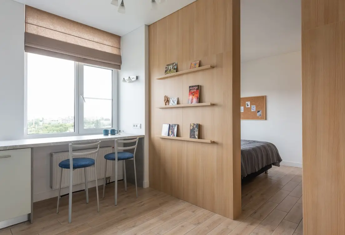 An apartment interior with an open wood bookshelf room divider separating a kitchen nook from the bedroom