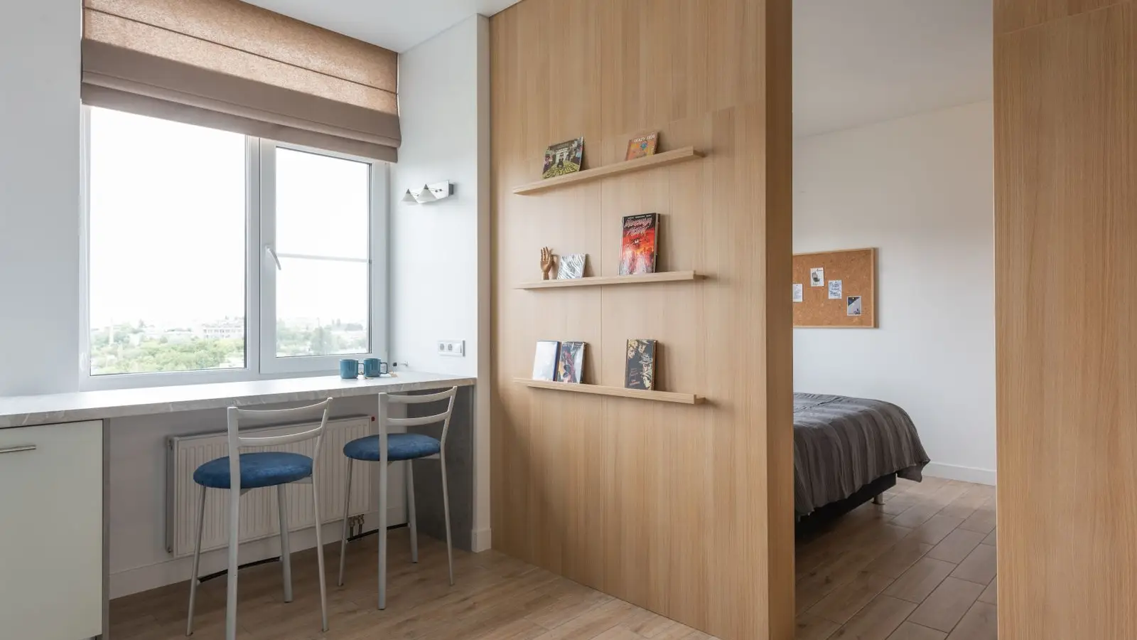 An apartment interior with an open wood bookshelf room divider separating a kitchen nook from the bedroom