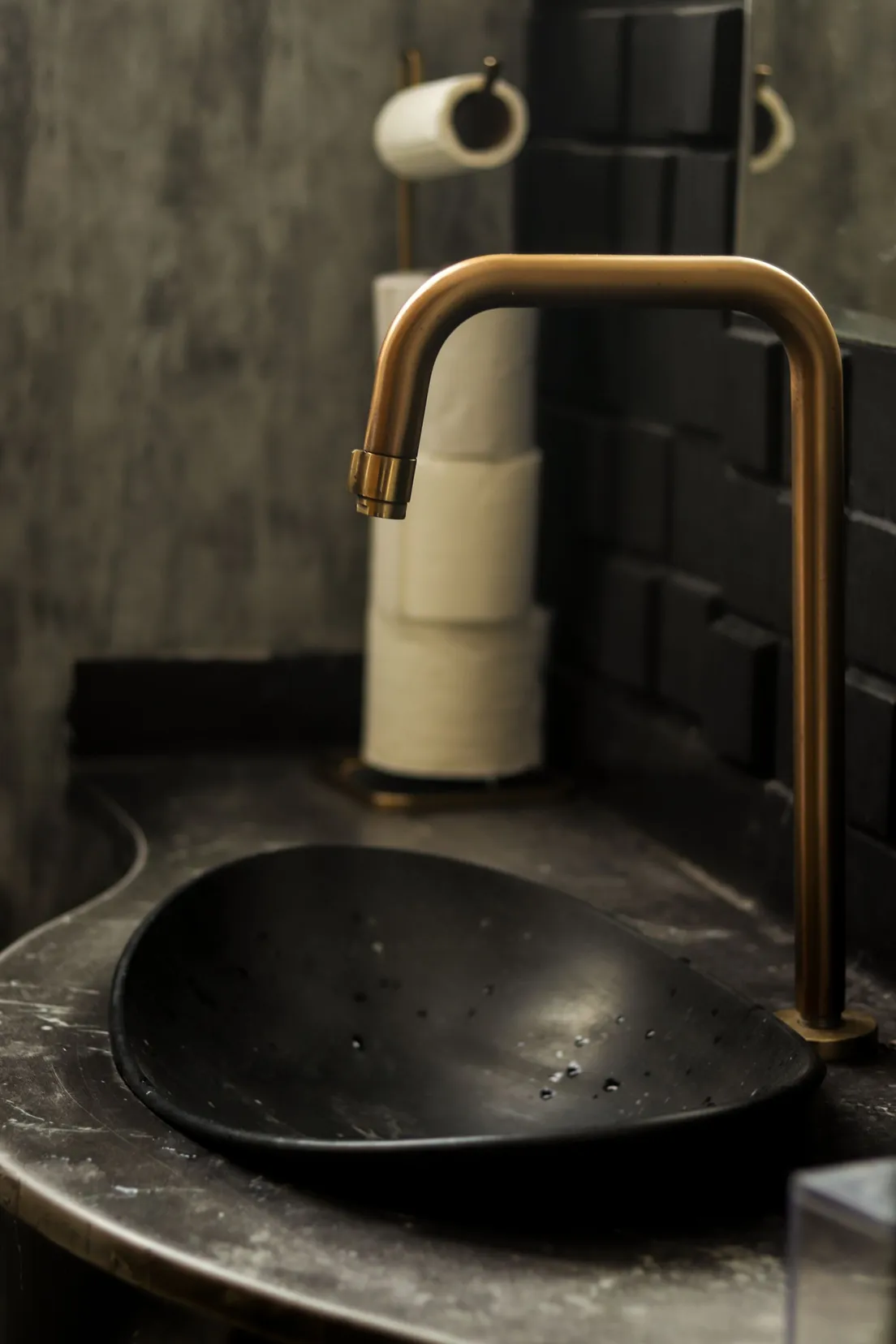 A close-up of a brass gooseneck faucet over a black stone sink basin, with a toilet paper holder in the background