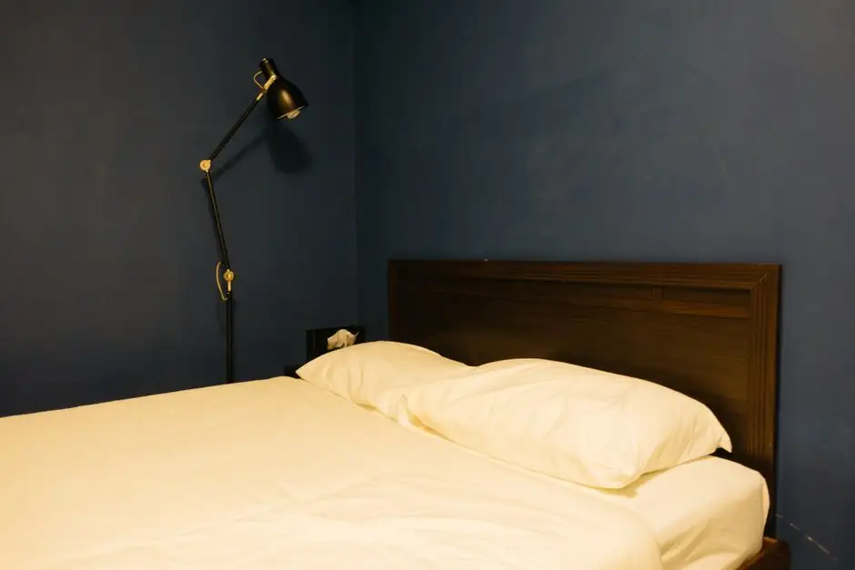 A minimalist bedroom with dark blue walls and warm lamp light, an example of a calming bedroom wall color for better sleep