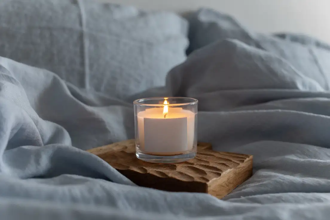 A lit candle in a glass holder on a carved wood tray resting on soft blue linen bedding