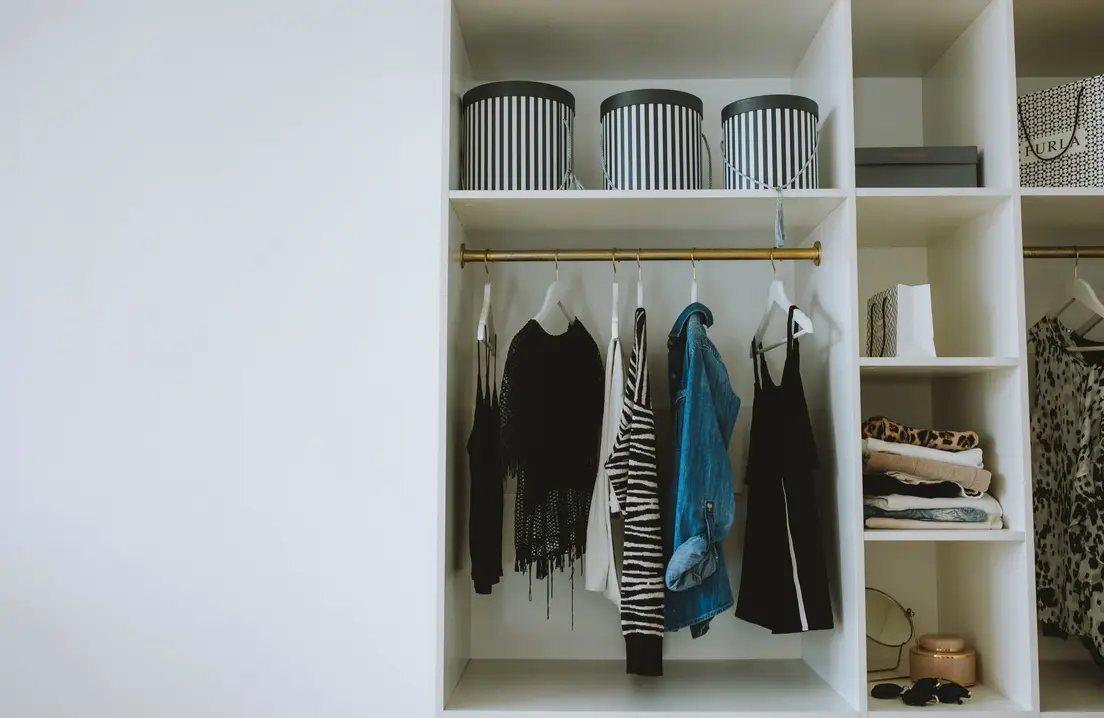 An organized small apartment closet with hanging clothes sorted by category and storage bins on the shelf above