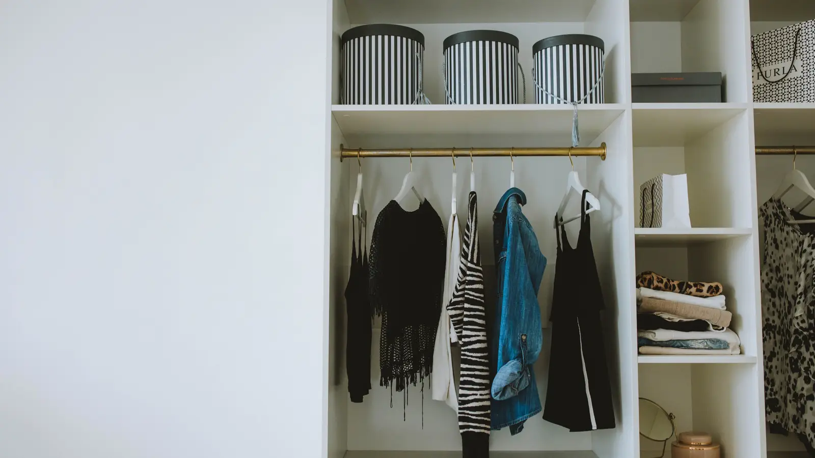 An organized small apartment closet with hanging clothes sorted by category and storage bins on the shelf above