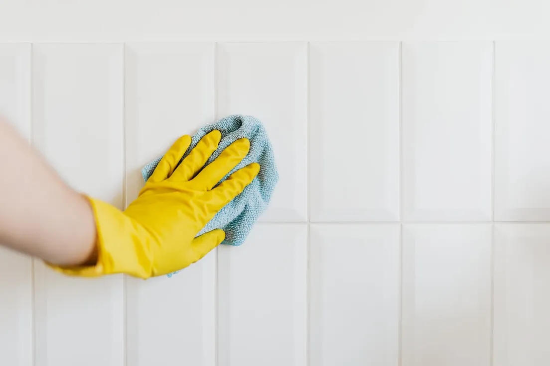 A gloved hand wiping a white tile wall clean with a cloth before installing a peel and stick backsplash