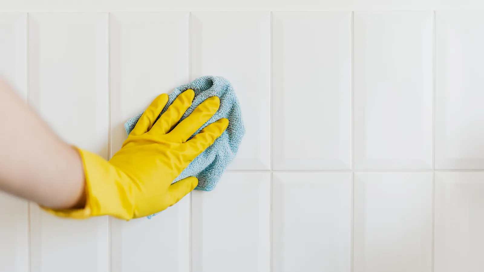A gloved hand wiping a white tile wall clean with a cloth before installing a peel and stick backsplash