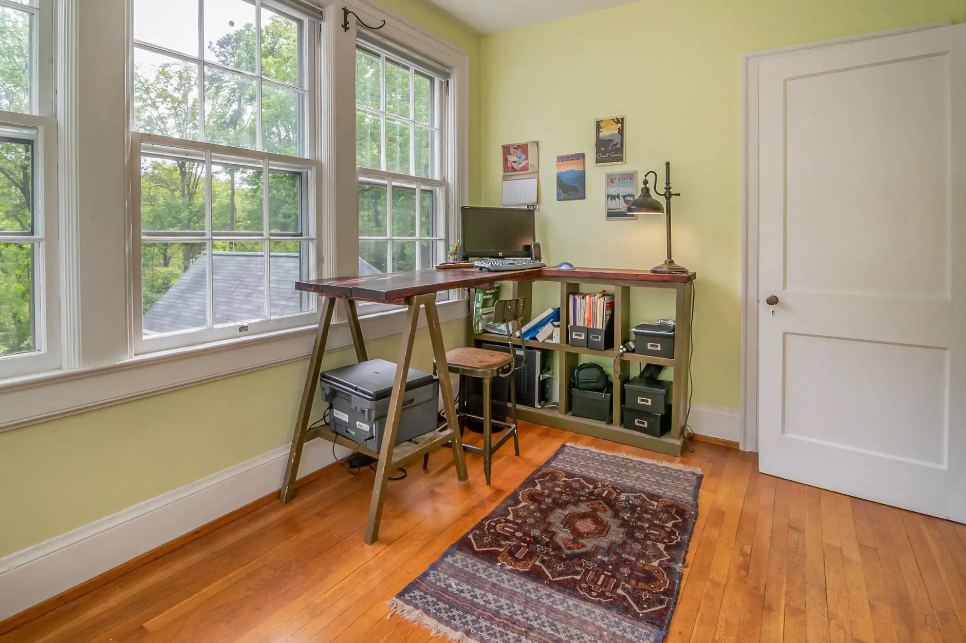 A compact trestle desk with shelving tucked into the corner of a room beside tall windows, an office in the living room corner