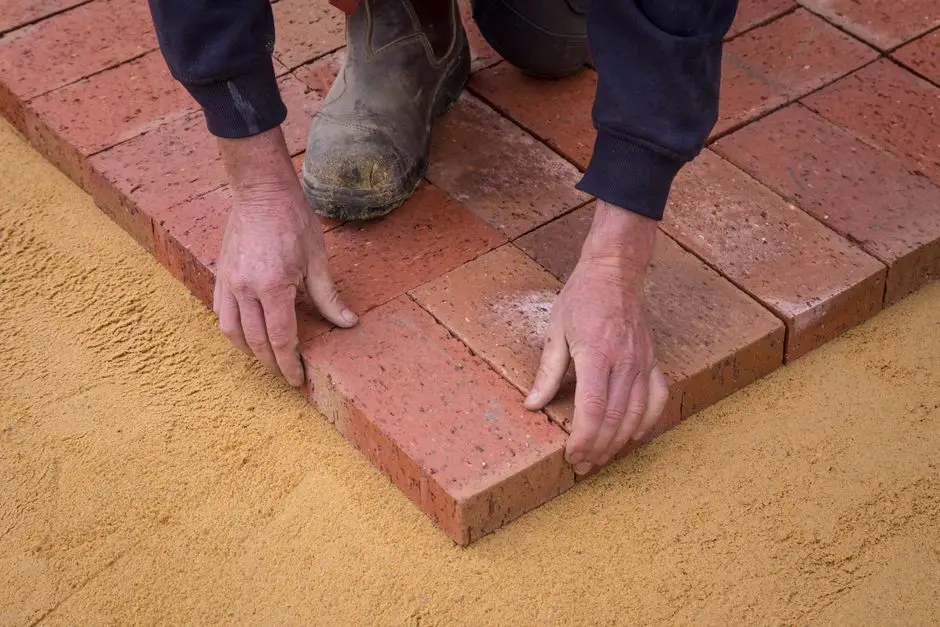 Hands laying brick pavers in sand, illustrating the labor cost difference between a porch and a patio