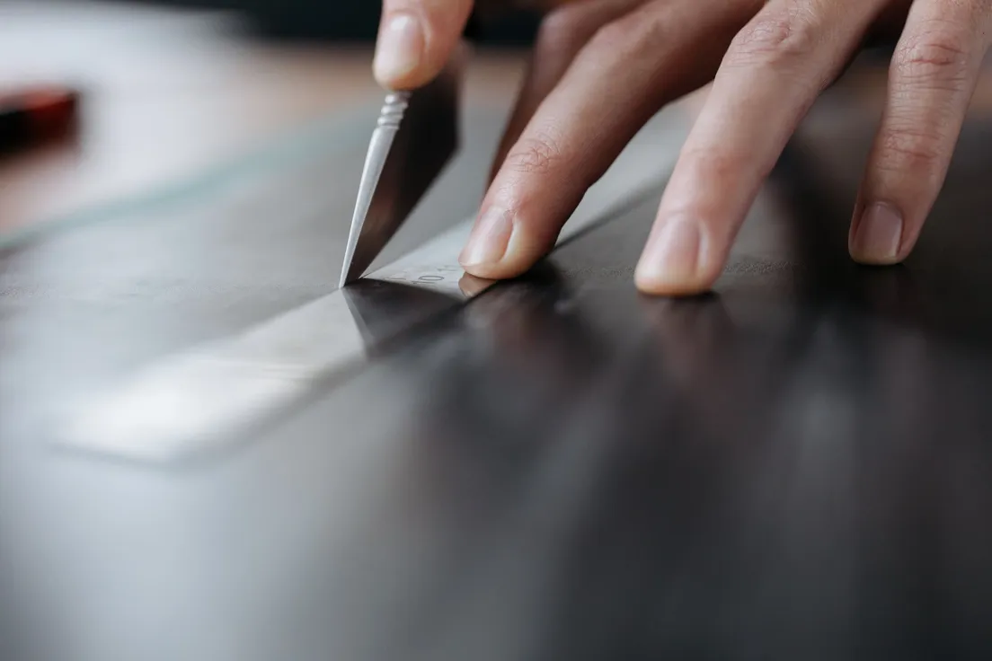 A hand using a utility knife and metal ruler to make a precise cut in a tile sheet