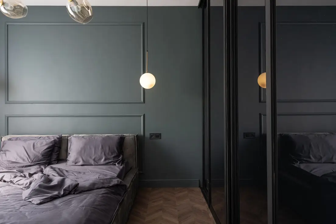 A deep slate-green bedroom accent wall with panel molding, a low grey bed, and a globe pendant light