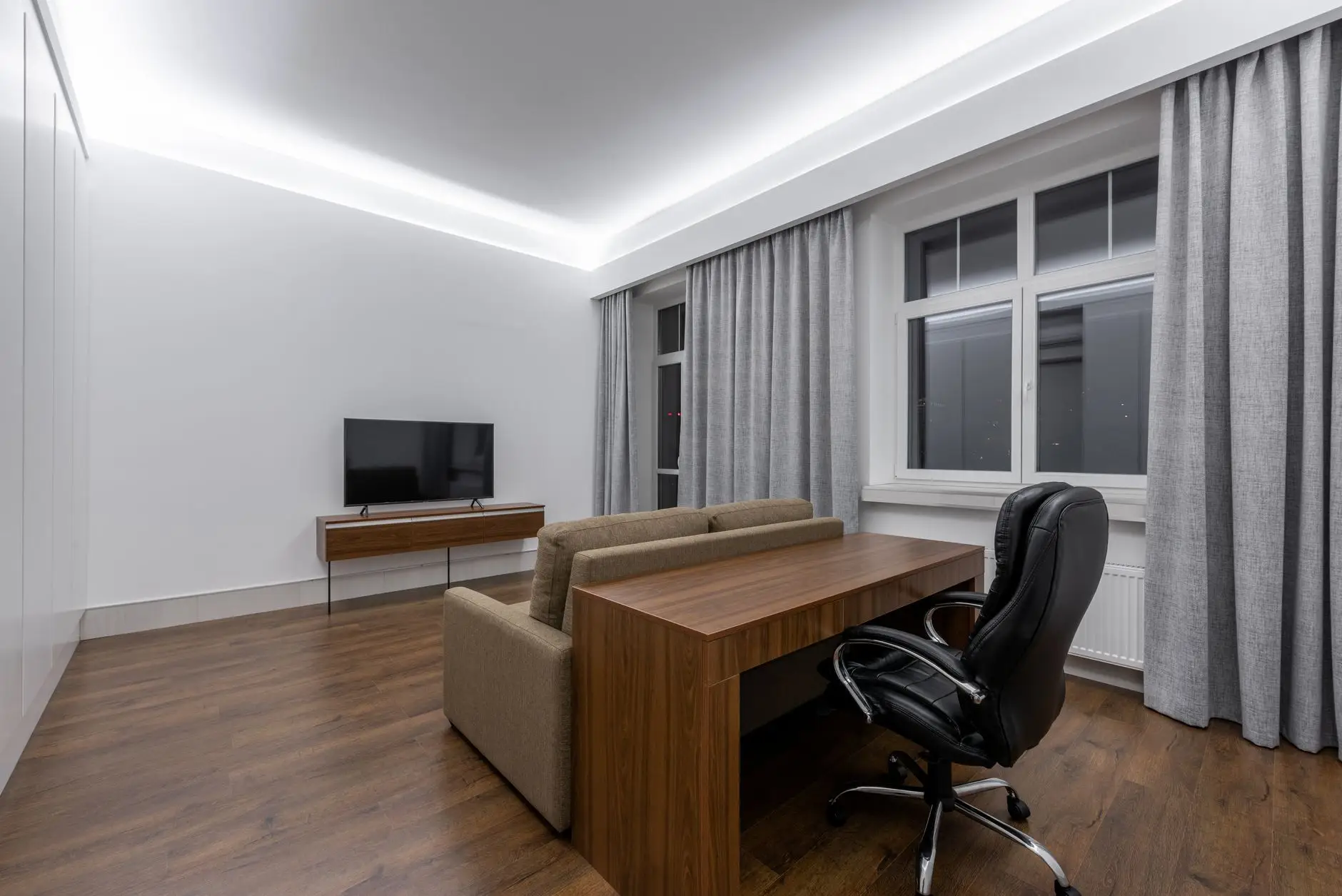 A wood desk placed directly behind a sofa with an office chair on the far side, a living room office layout facing away from the TV