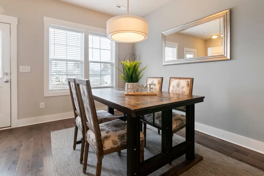 A large framed mirror on the wall of a small dining area above a wood dining table with upholstered chairs and a drum pendant light