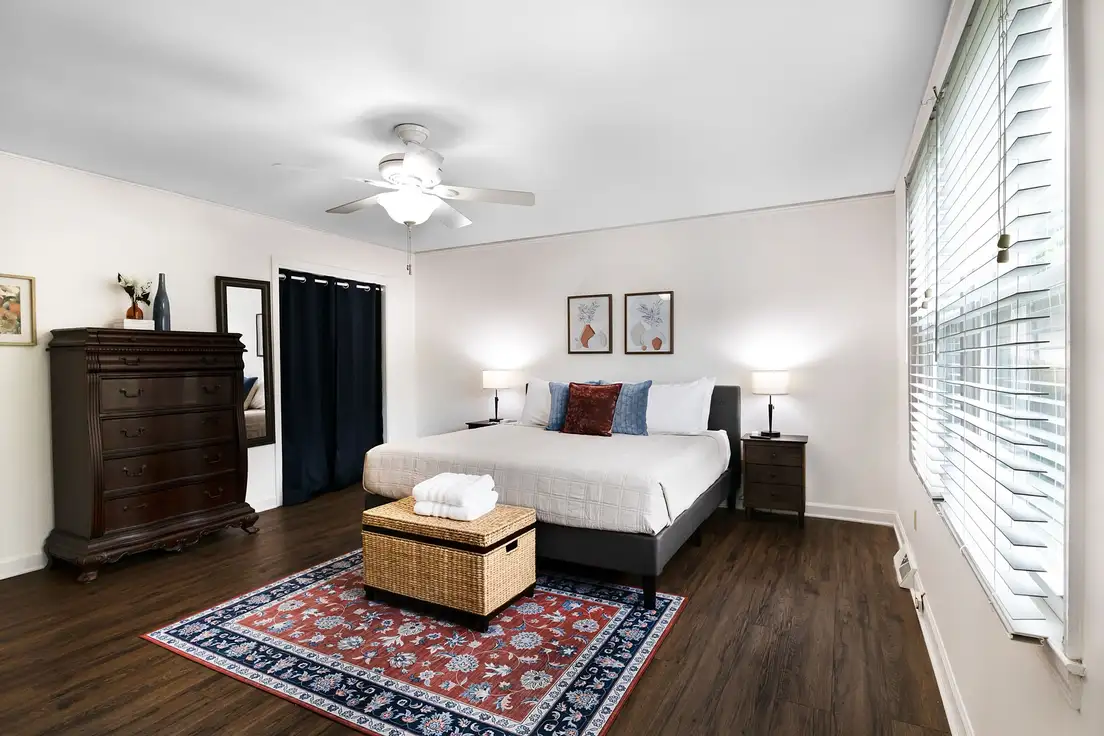 A bedroom with a tall wood dresser, a wicker storage trunk at the foot of the bed, and a patterned area rug