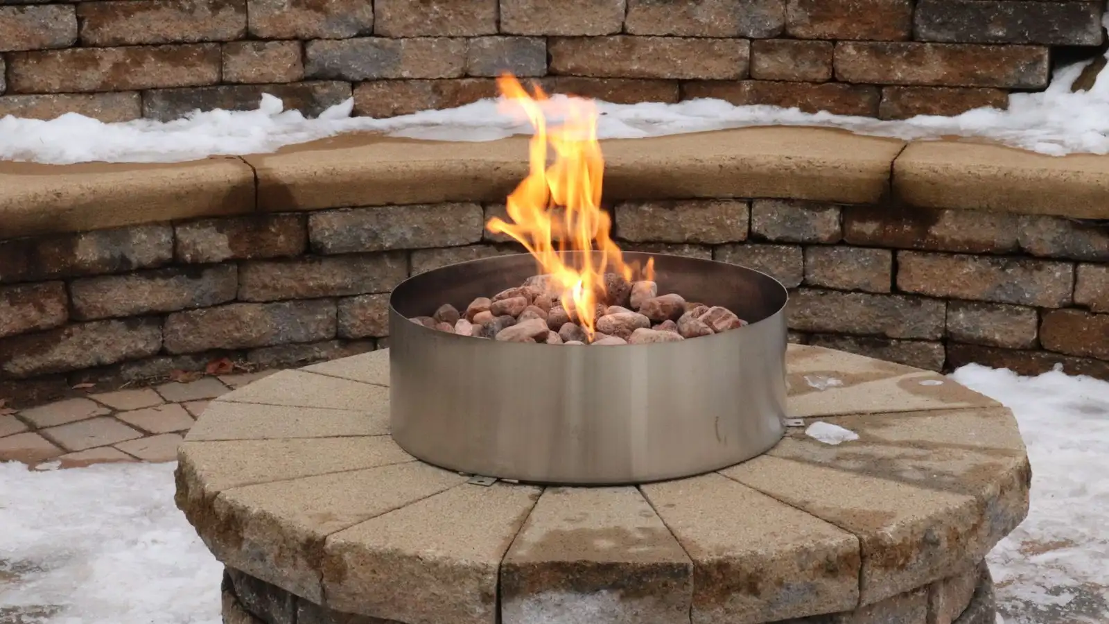 A stainless steel fire pit ring insert set into a stone paver surround with a curved stone seat wall