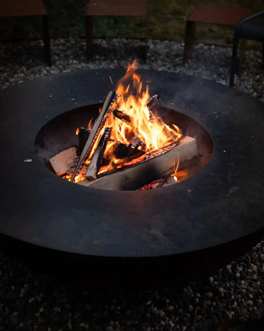 A steel fire pit bowl burning on a pea gravel patio surface