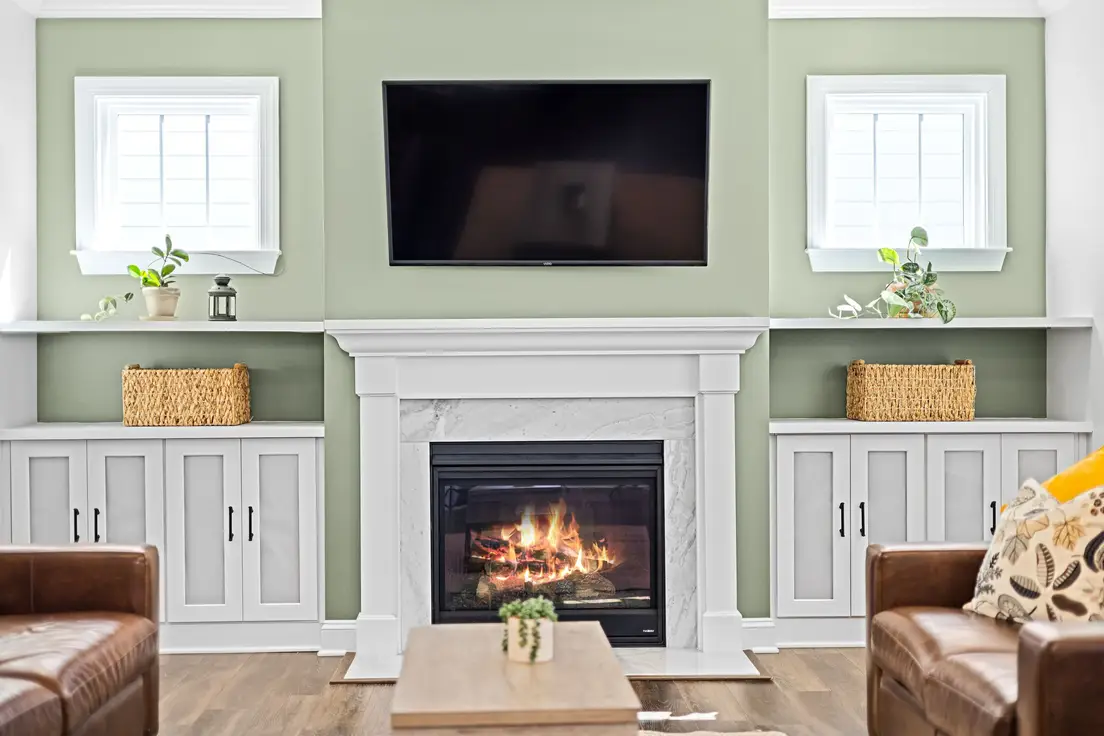 A TV mounted above a white fireplace mantel on a sage green wall flanked by built-in cabinets