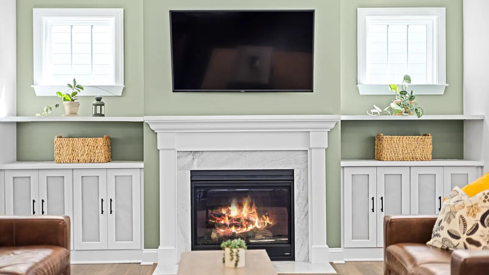 A TV mounted above a white fireplace mantel on a sage green wall flanked by built-in cabinets