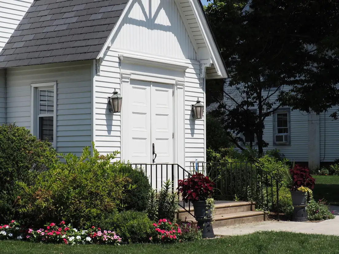 A white cottage entry with a flower bed of pink impatiens, red potted plants, and shrubs along the front steps