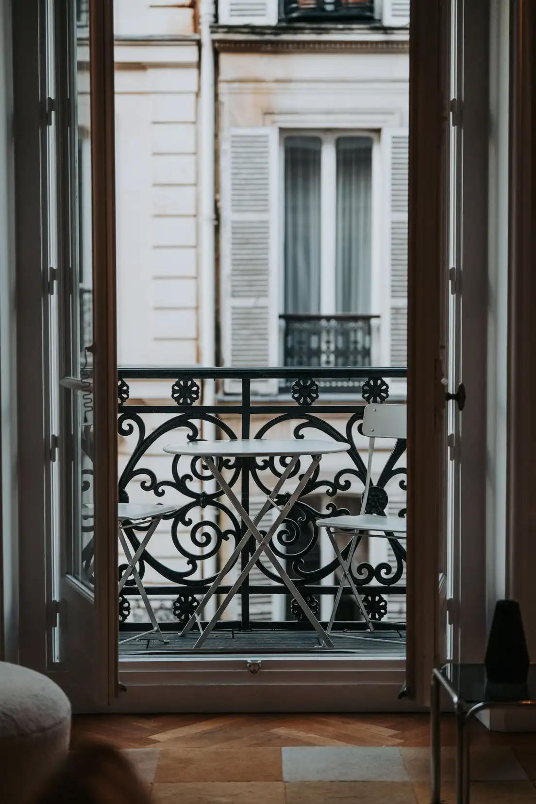 A small round bistro table with two folding chairs seen through open French doors onto a Paris apartment balcony