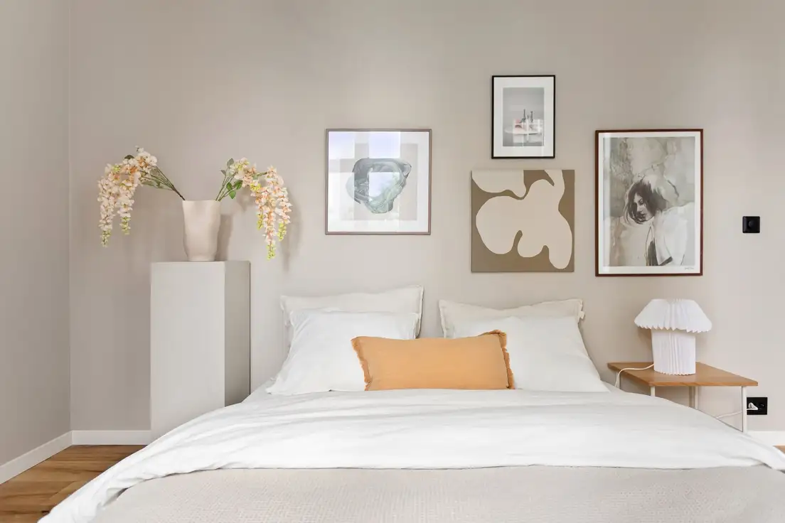 A greige bedroom wall with a small gallery of framed prints above a bed with white bedding and a peach pillow