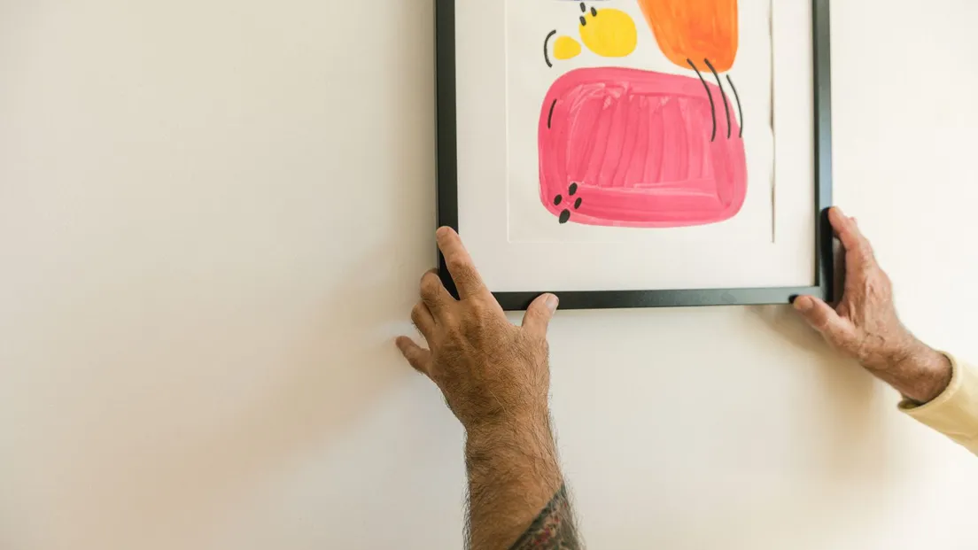Hands hanging a bright abstract print in a black frame on a white wall for a no-nail gallery wall