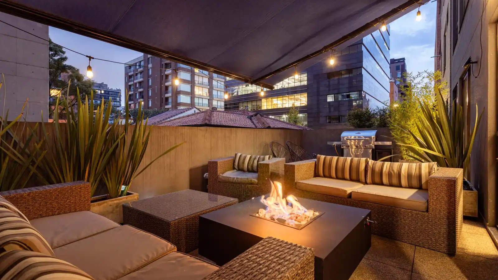 A lit rectangular propane fire table used as a coffee table on a small patio with wicker sofas and string lights