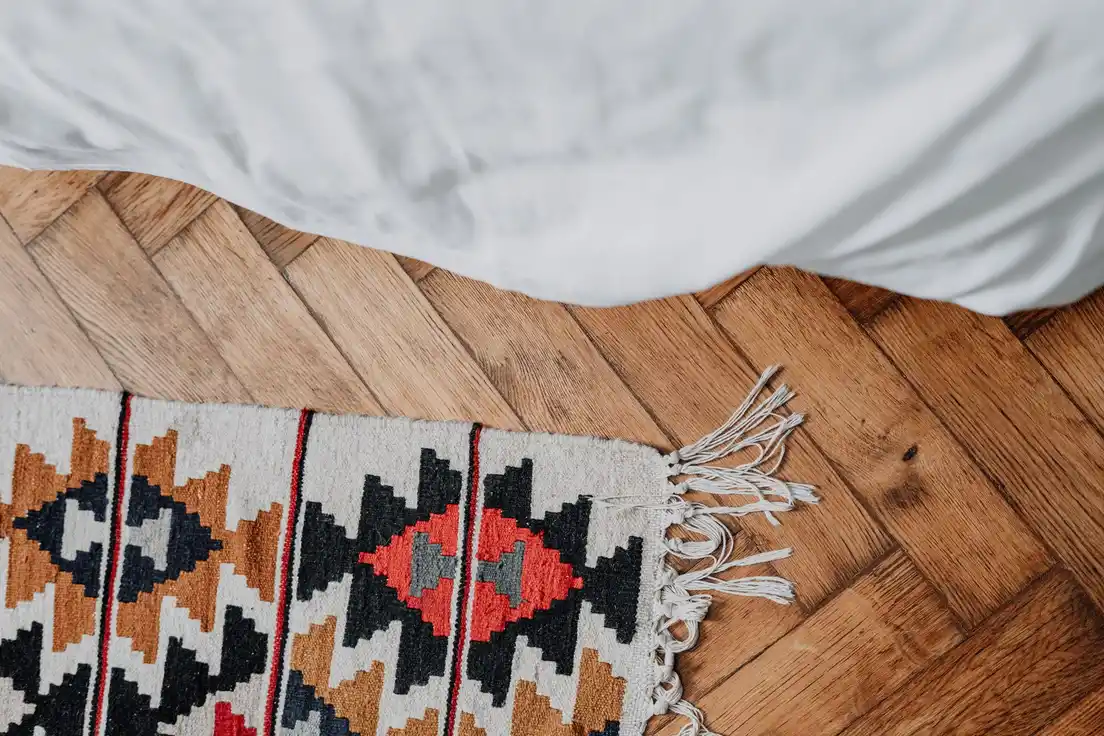 A geometric flatweave kilim rug with fringe on a herringbone wood floor beside a bed