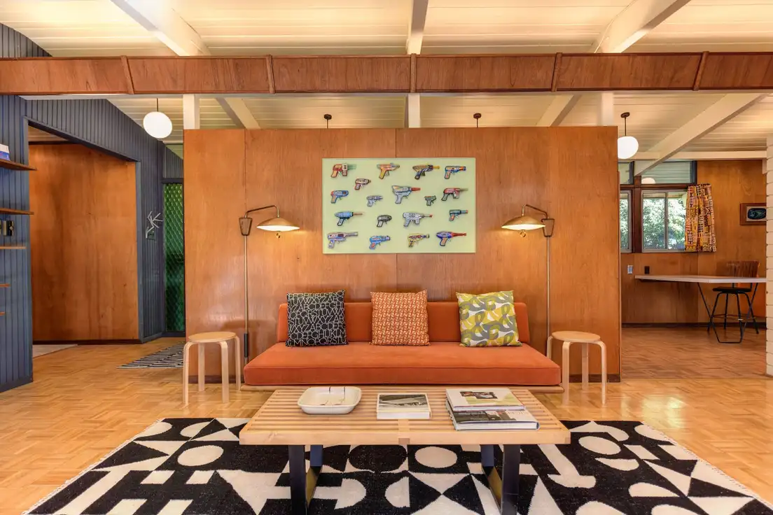 Mid-century modern lighting in a ranch living room: white globe pendants under a beamed ceiling and brass swing-arm wall sconces beside an orange sofa