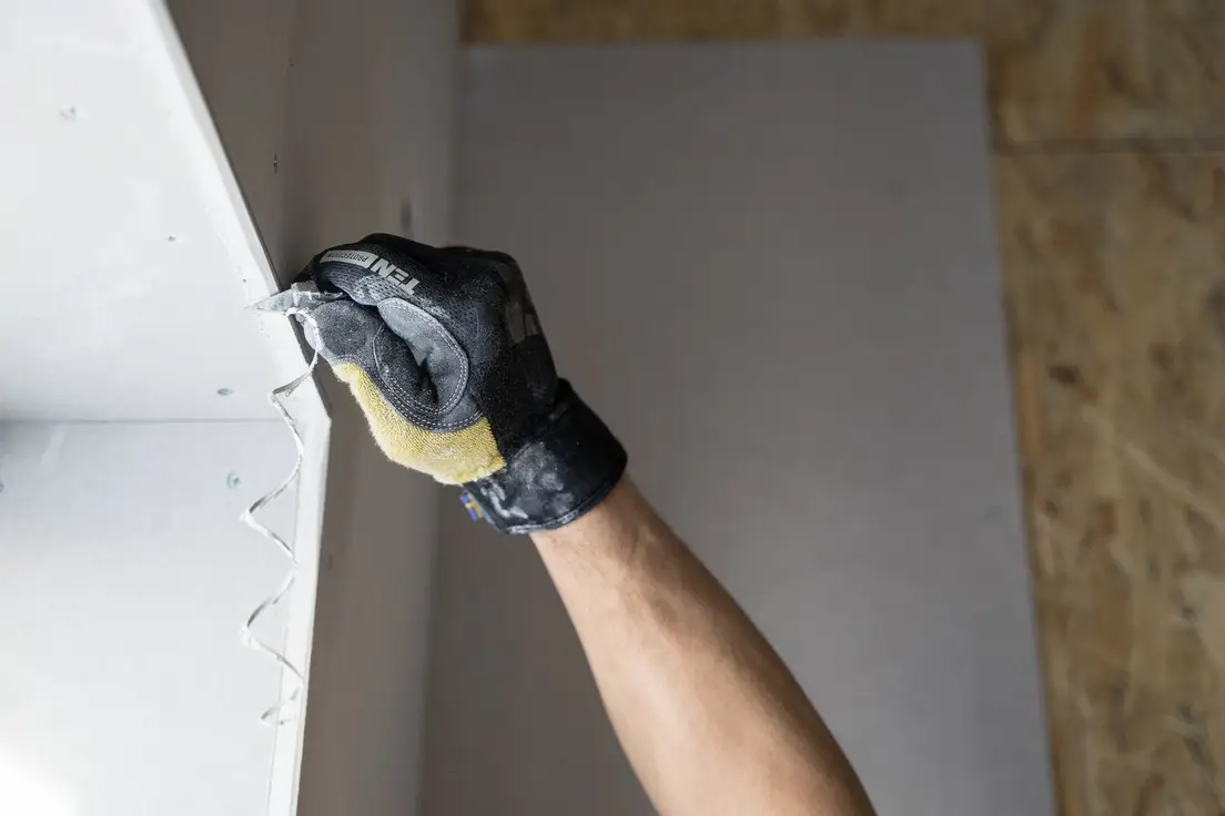A gloved hand cutting a drywall panel edge during installation of a temporary wall