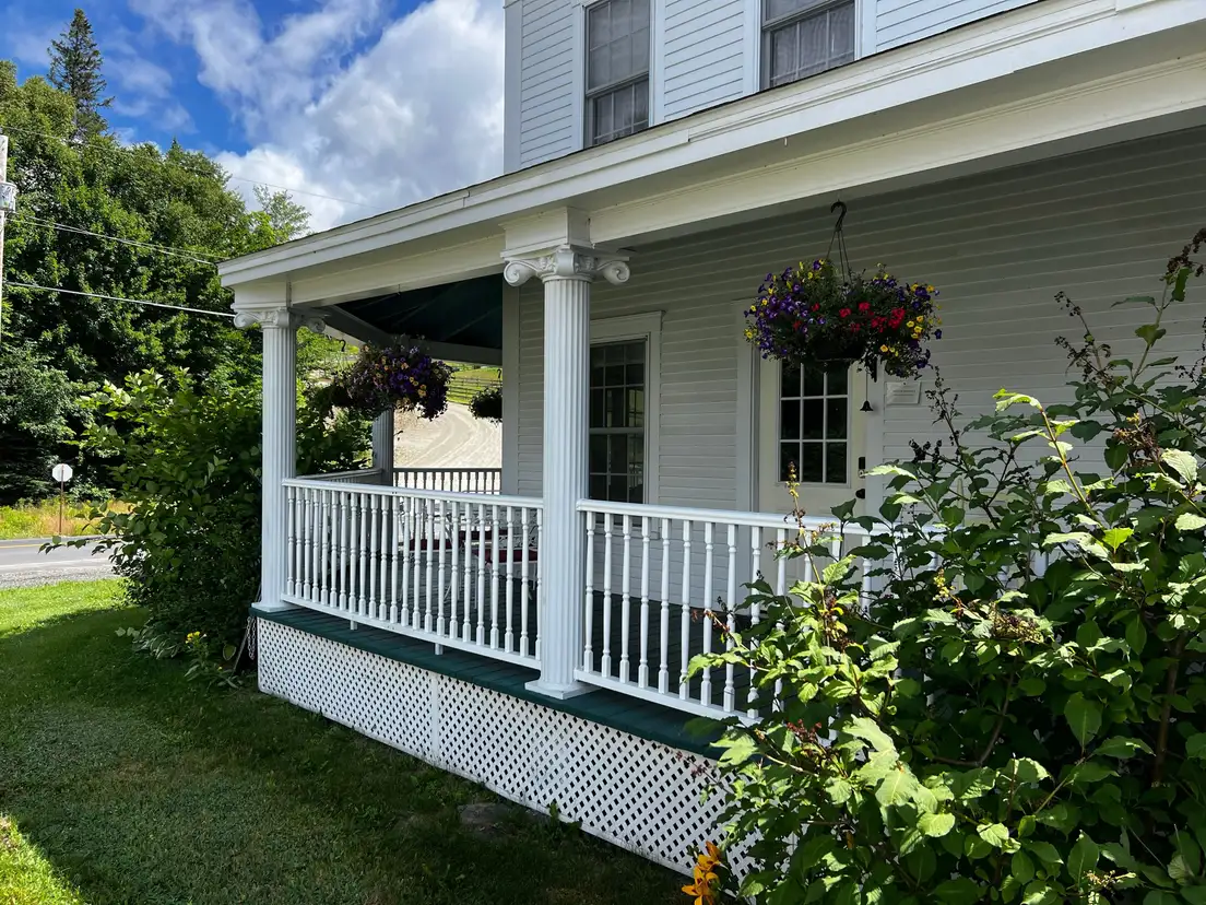 A white wraparound porch with turned columns, white railings, and hanging baskets of purple flowers