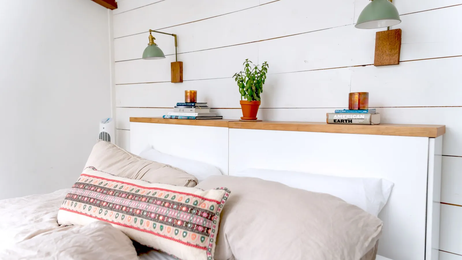 A white floating shelf headboard with wall sconces, books, and a small potted plant above a bed