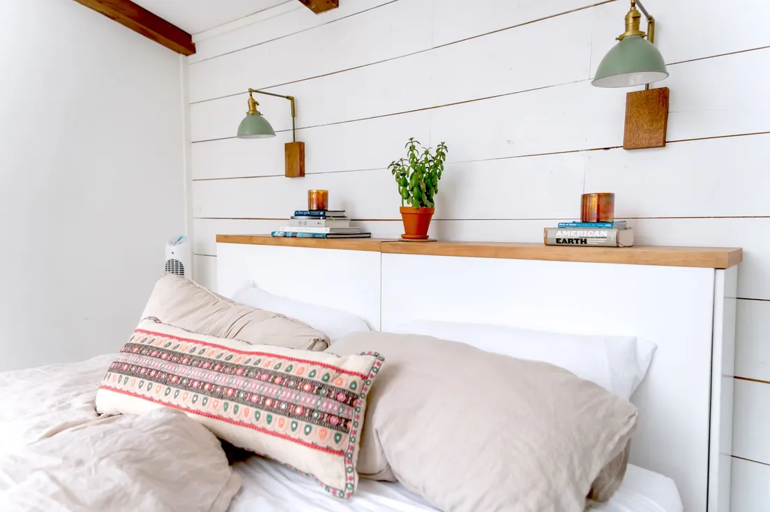 A white floating shelf headboard with wall sconces, books, and a small potted plant above a bed