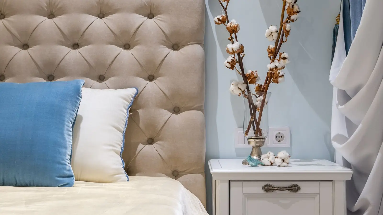 An upholstered headboard with cotton stems in a vase on the nightstand beside it
