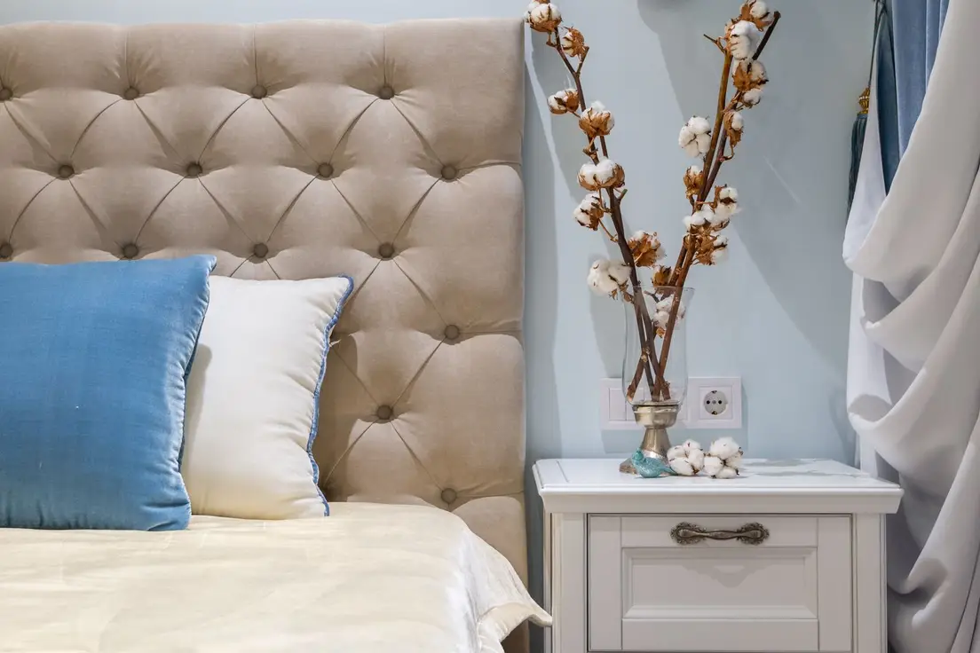 An upholstered headboard with cotton stems in a vase on the nightstand beside it