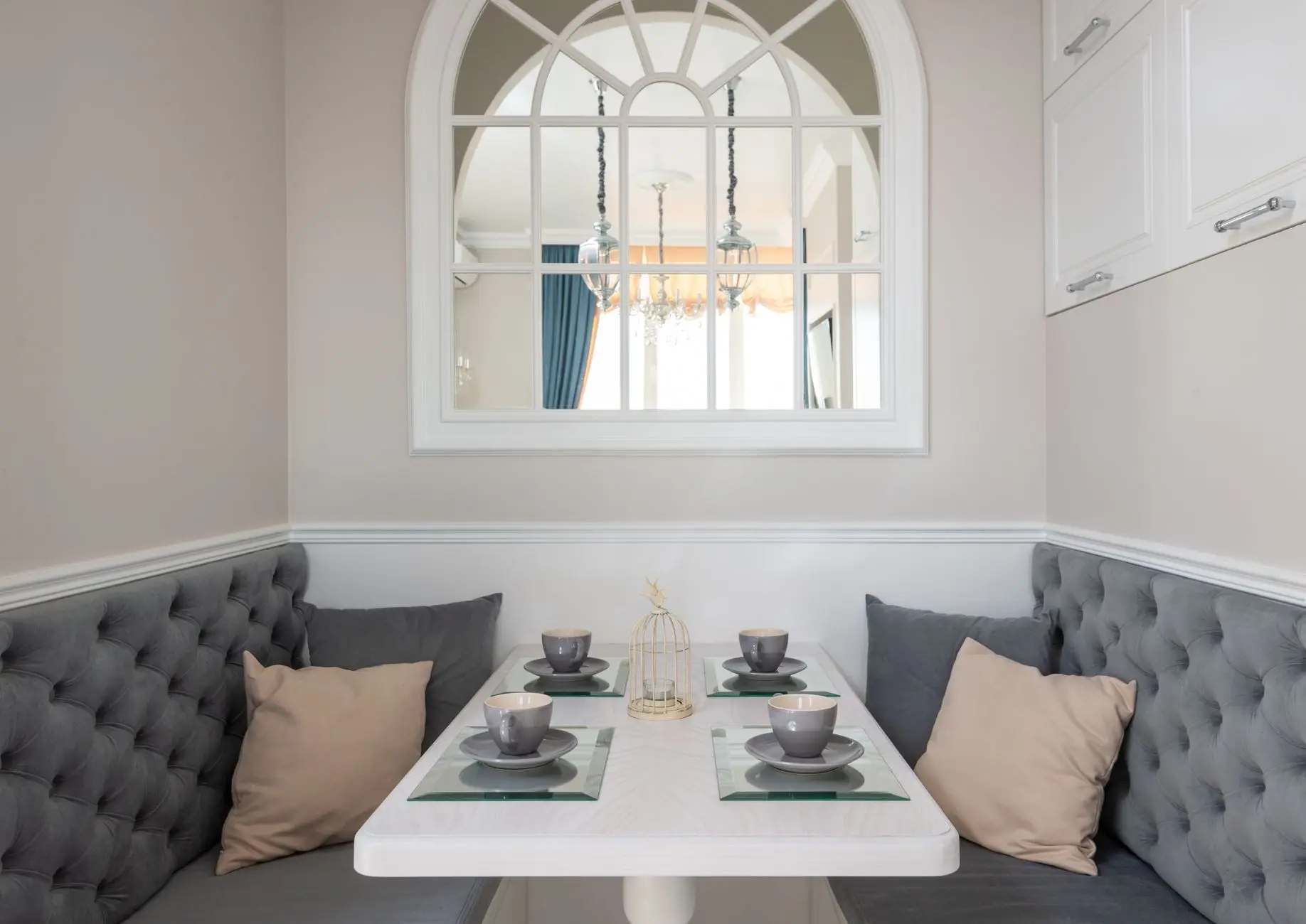 A small kitchen nook with a curved gray tufted banquette bench, a white table, and an arched window