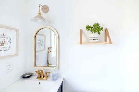 A small bathroom with a round mirror, wall shelf, and potted plant in warm natural light